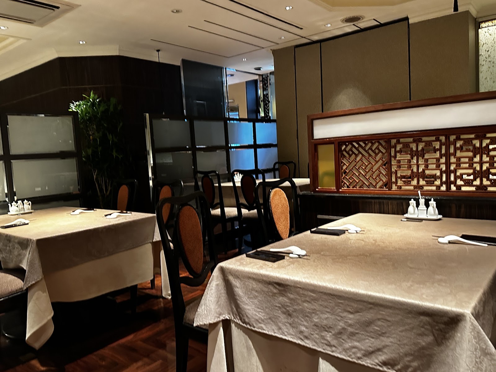 白いレンゲと箸、調味料のポットがセットされたシャンパン色のダマスク織のテーブル、籐の背の黒い椅子、彫刻を施した紫檀の格子衝立、曇りガラスの衝立、観葉植物、ヘリンボーンの床のダイニング。