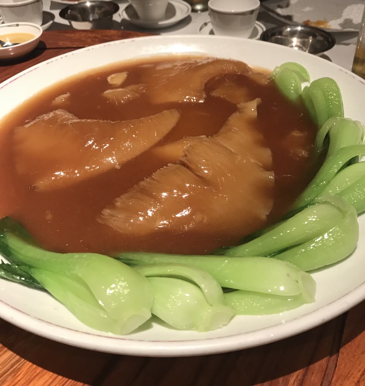 赤い縁の白い大皿に、照りのある茶色のソースで煮込んだ姿ふかひれとそれを囲む青梗菜。木のテーブル。
