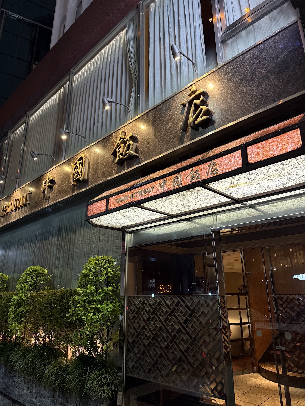 夜の店構え。スポットライトに照らされた黒い御影石の壁に金色の浮き彫りの「中國飯店」の文字、珊瑚色に光るガラスの庇に「CHINESE RESTAURANT 中國飯店」、網代格子の衝立が見えるガラス扉、照明の当たった植え込み。