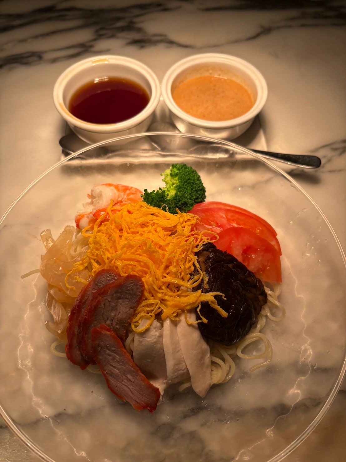 大理石のテーブルの上の冷麺。錦糸卵、チャーシュー、海老、くらげ、トマト、ブロッコリーを載せ、奥に二種のタレの器。