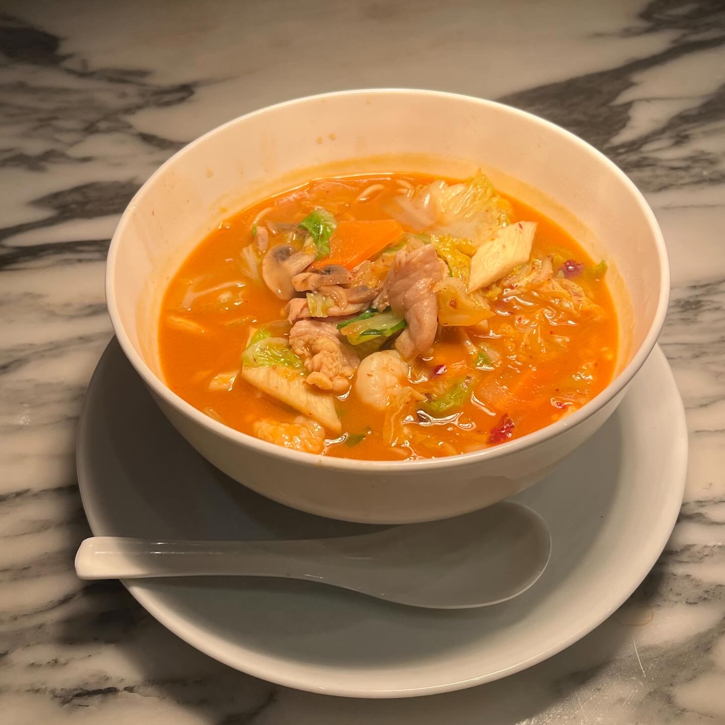 大理石のテーブルの上の白い器。赤いスープに麺、豚肉、白菜、海老。白いレンゲ。